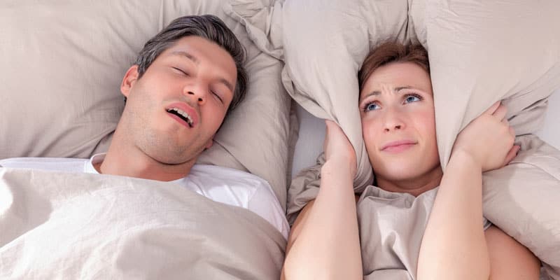 A man snoring with his mouth open next to his wife who is covering her ears and cannot sleep